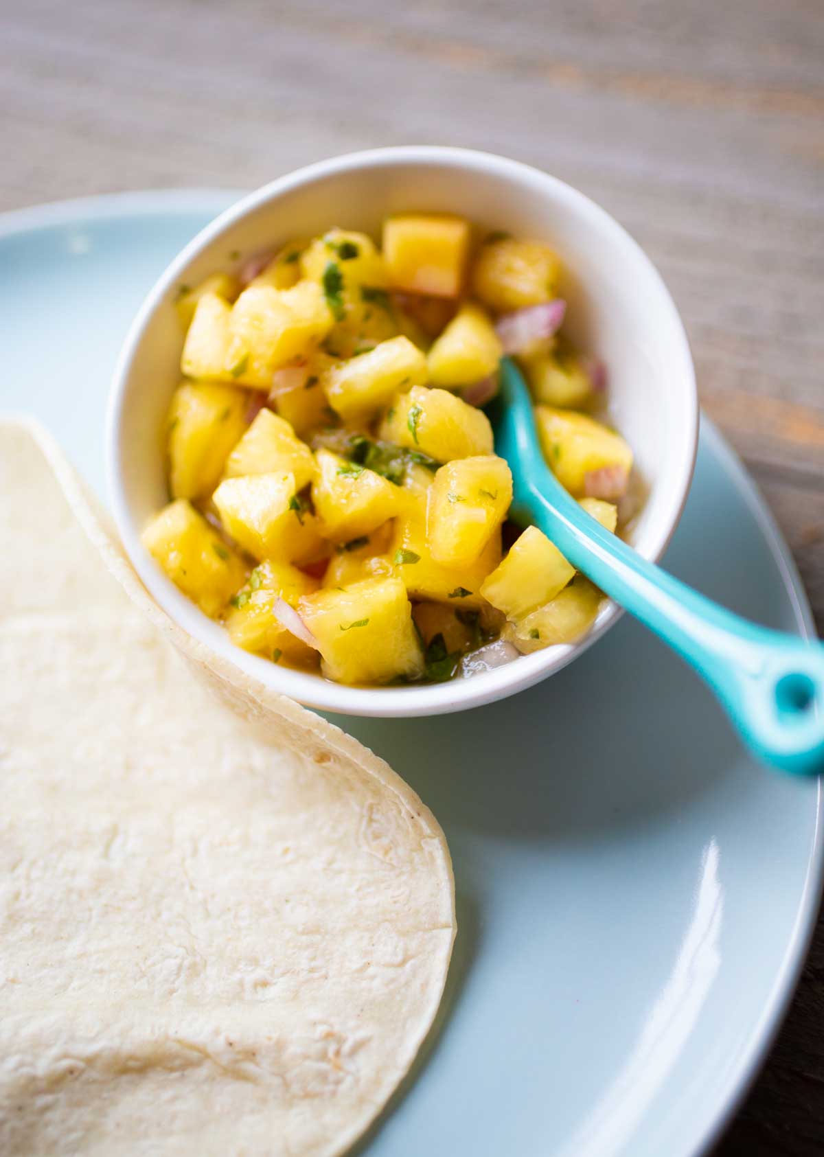 A small white bowl filled with pineapple salsa and a blue spoon.