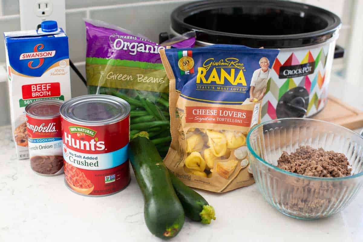 The ingredients to make a tomato based tortellini soup are on the counter next to a crockpot.