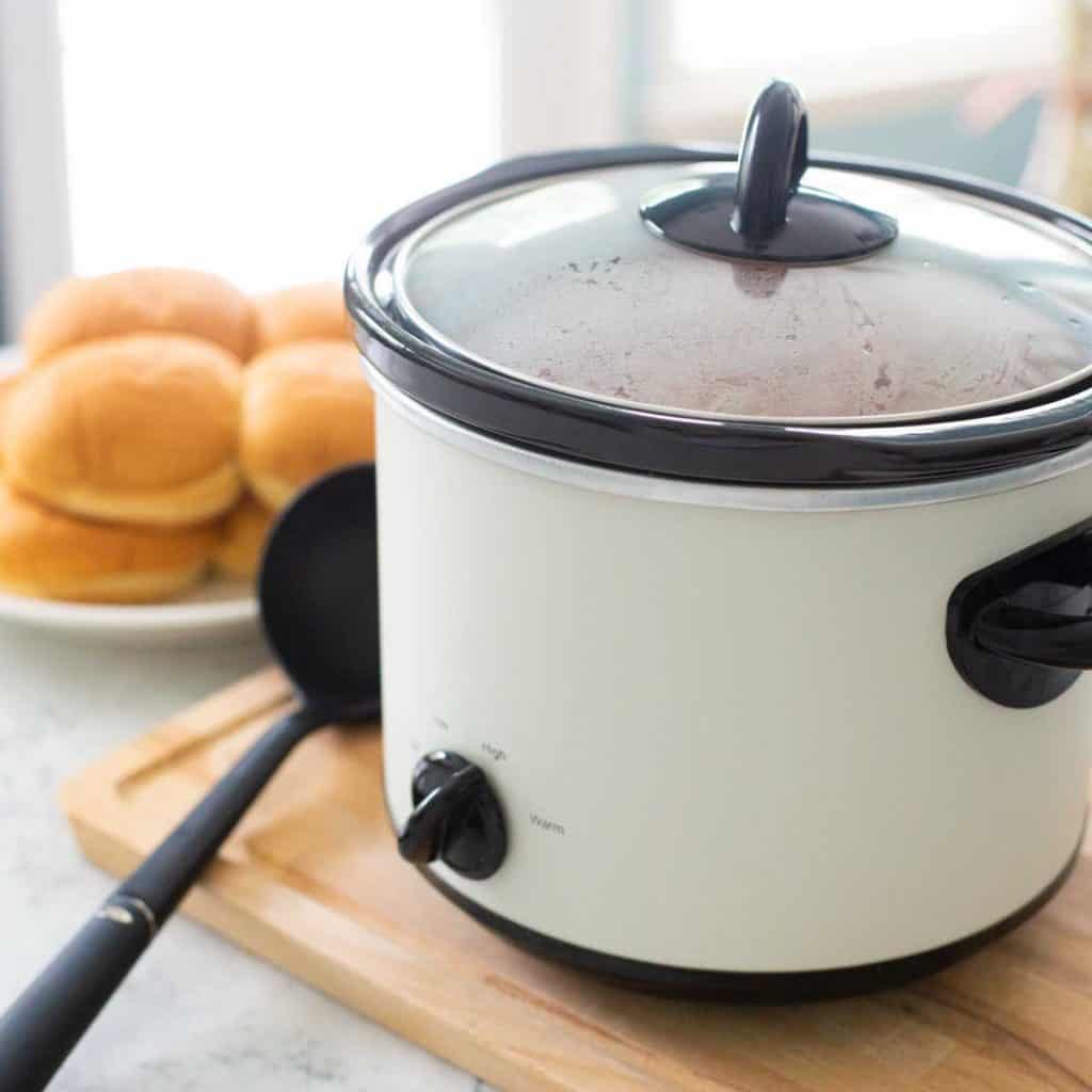 A slowcooker sits on a wooden cutting board. There's a ladle and a plate full of sandwich buns sitting next to it.