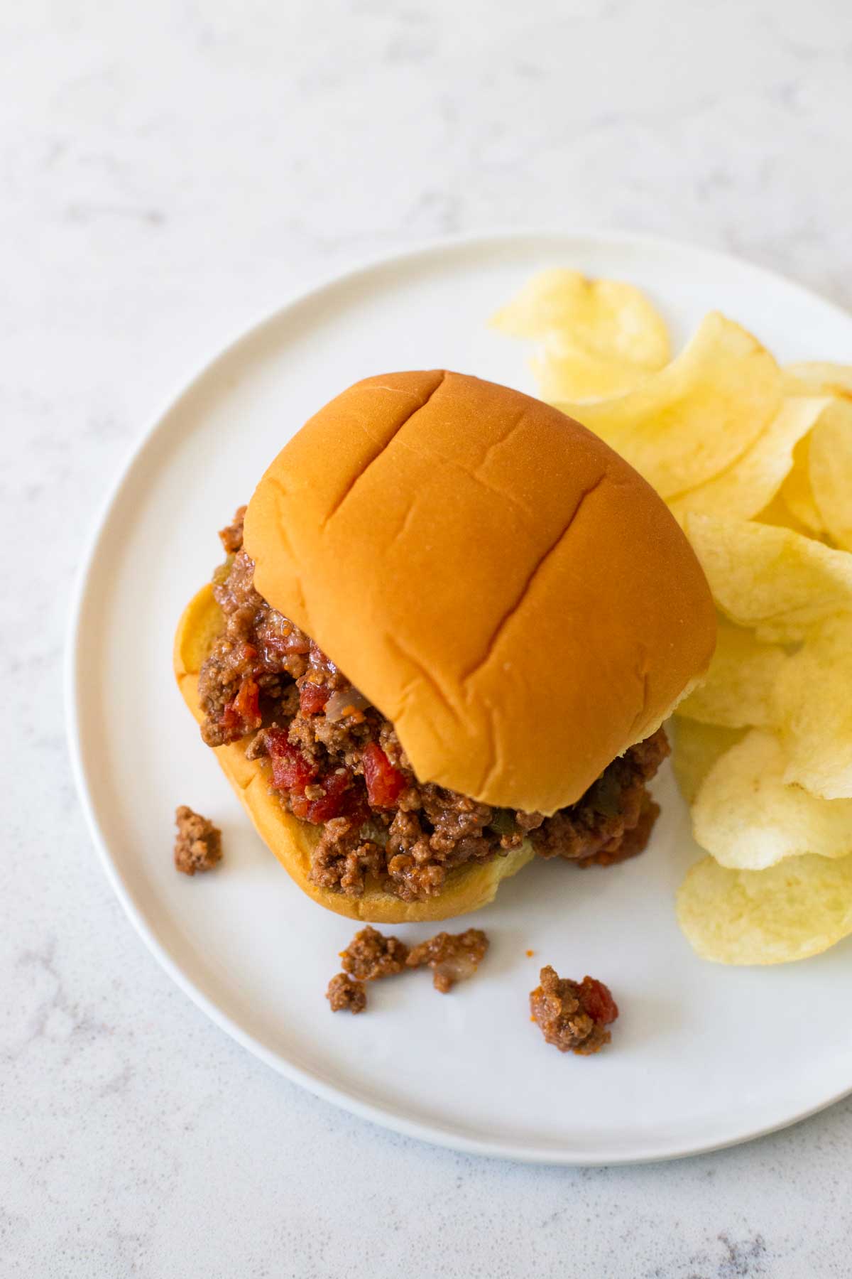 A sloppy joe sandwich with filling falling onto the plate has a pile of potato chips next to it.