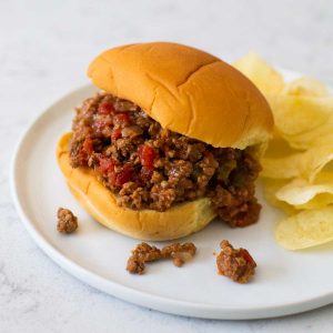 A sloppy joe sandwich on a soft bun has some filling dripping onto the plate and potato chips on the side.