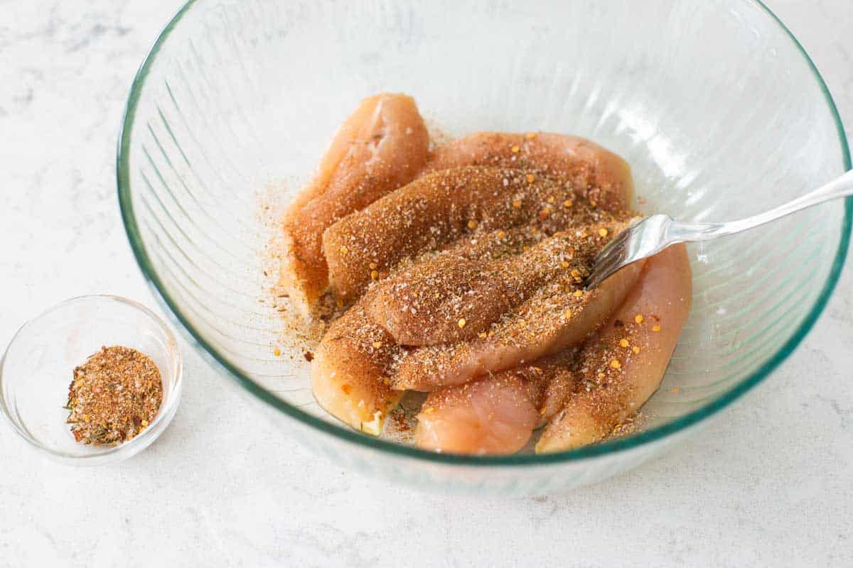 A mixing bowl filled with chicken tenderloins have been sprinkled with a jerk seasoning blend. The bowl of seasoning is on the side.