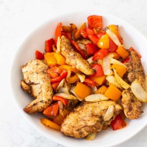A bowl of cooked jerk chicken with red and yellow bell peppers and onions.