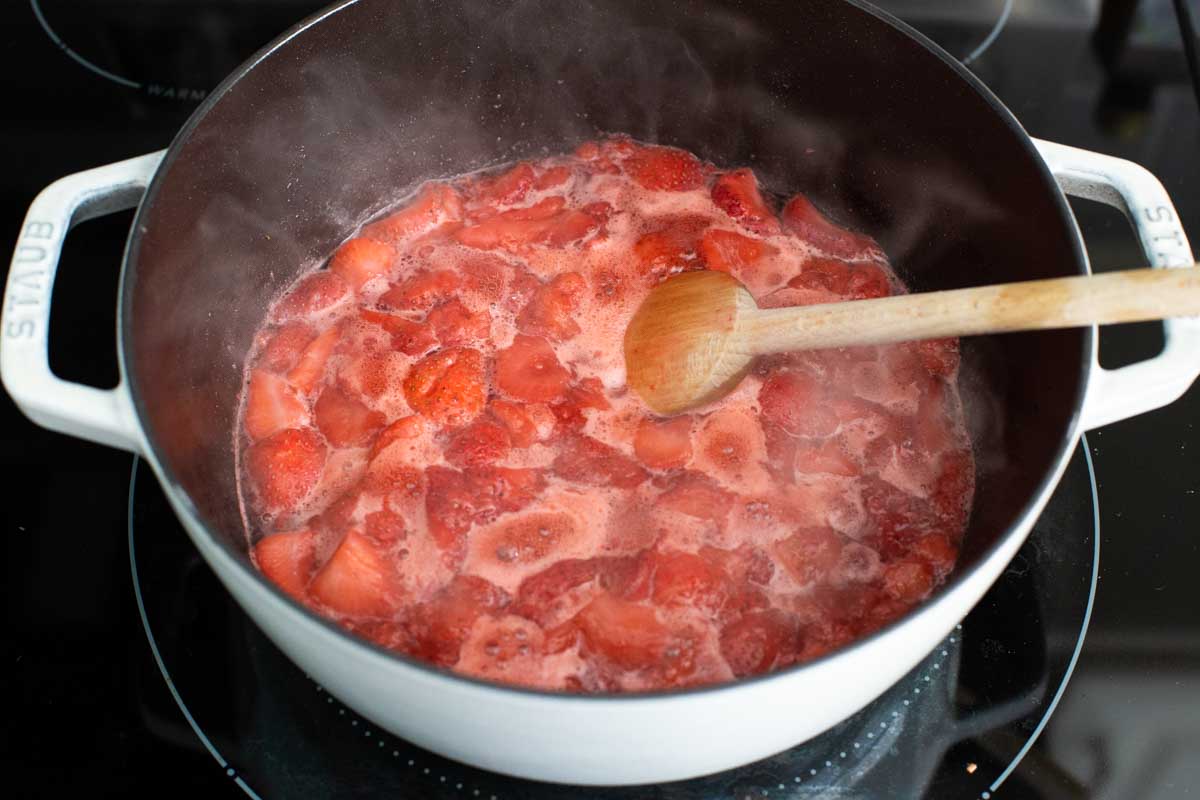 The strawberries and sugar are boiling in a large pot with a wooden spoon.