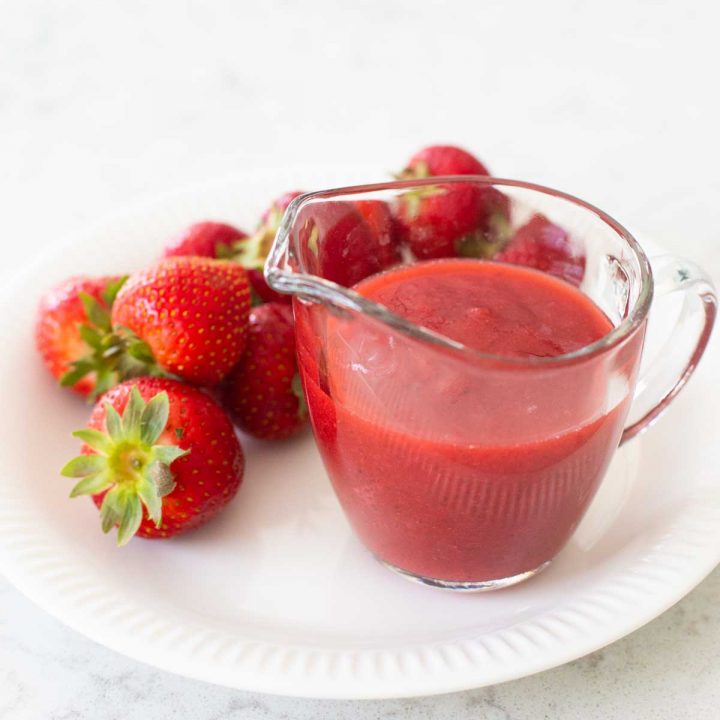 A pitcher of strawberry sauce sits on a plate next to fresh strawberries.
