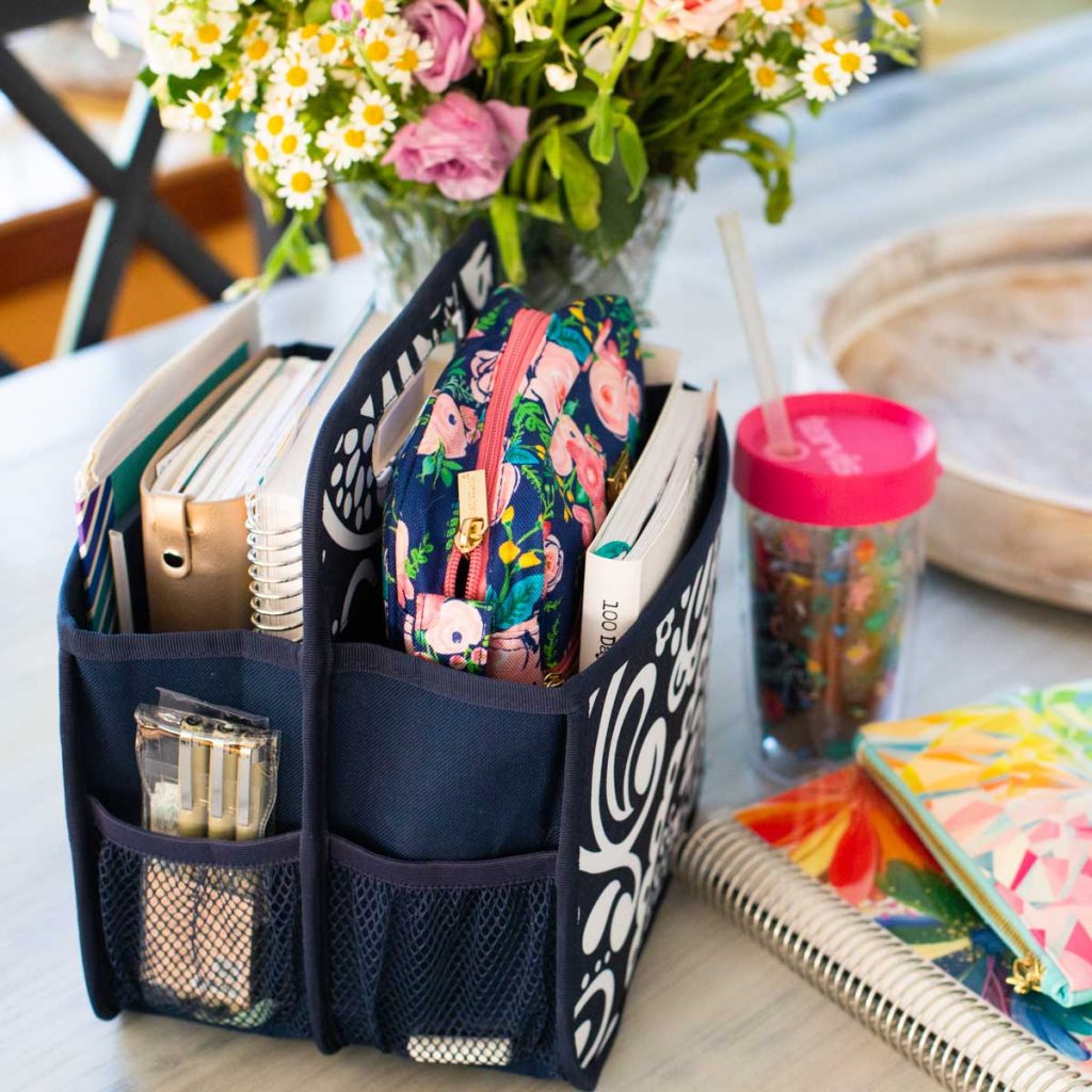 A basket of reading material is on a table with iced coffee, a planner, and fresh flowers.