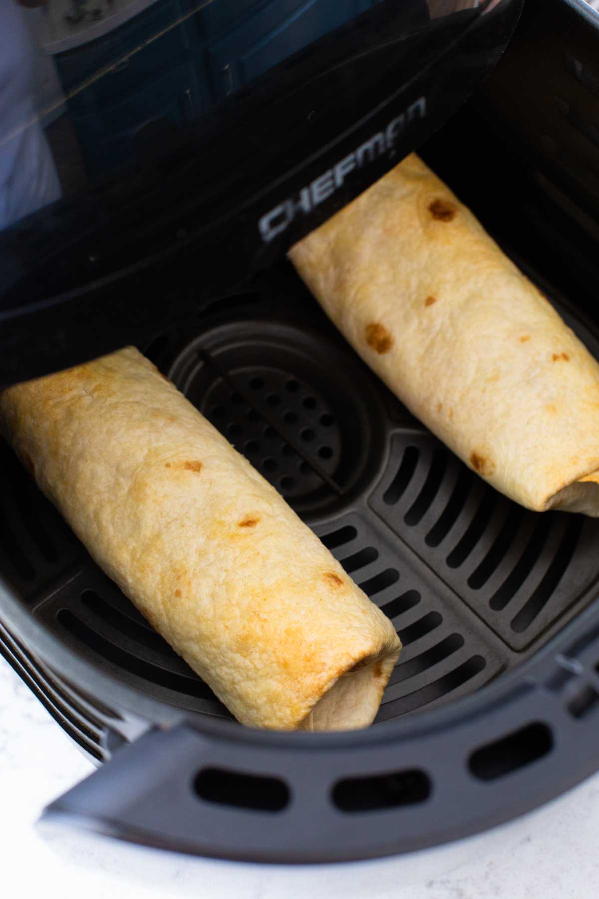 Two chimichangas cook in an air fryer basket.