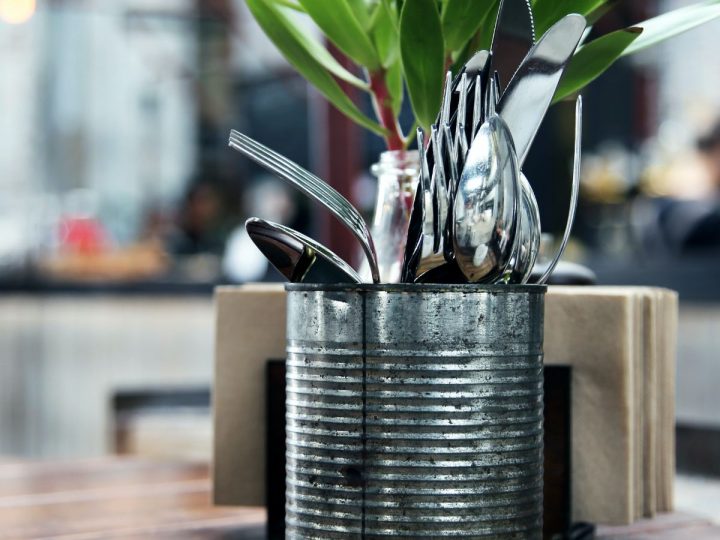 A metal can holds eating untensils on a table next to a plant.