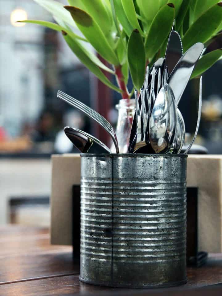 A metal can holds eating untensils on a table next to a plant.