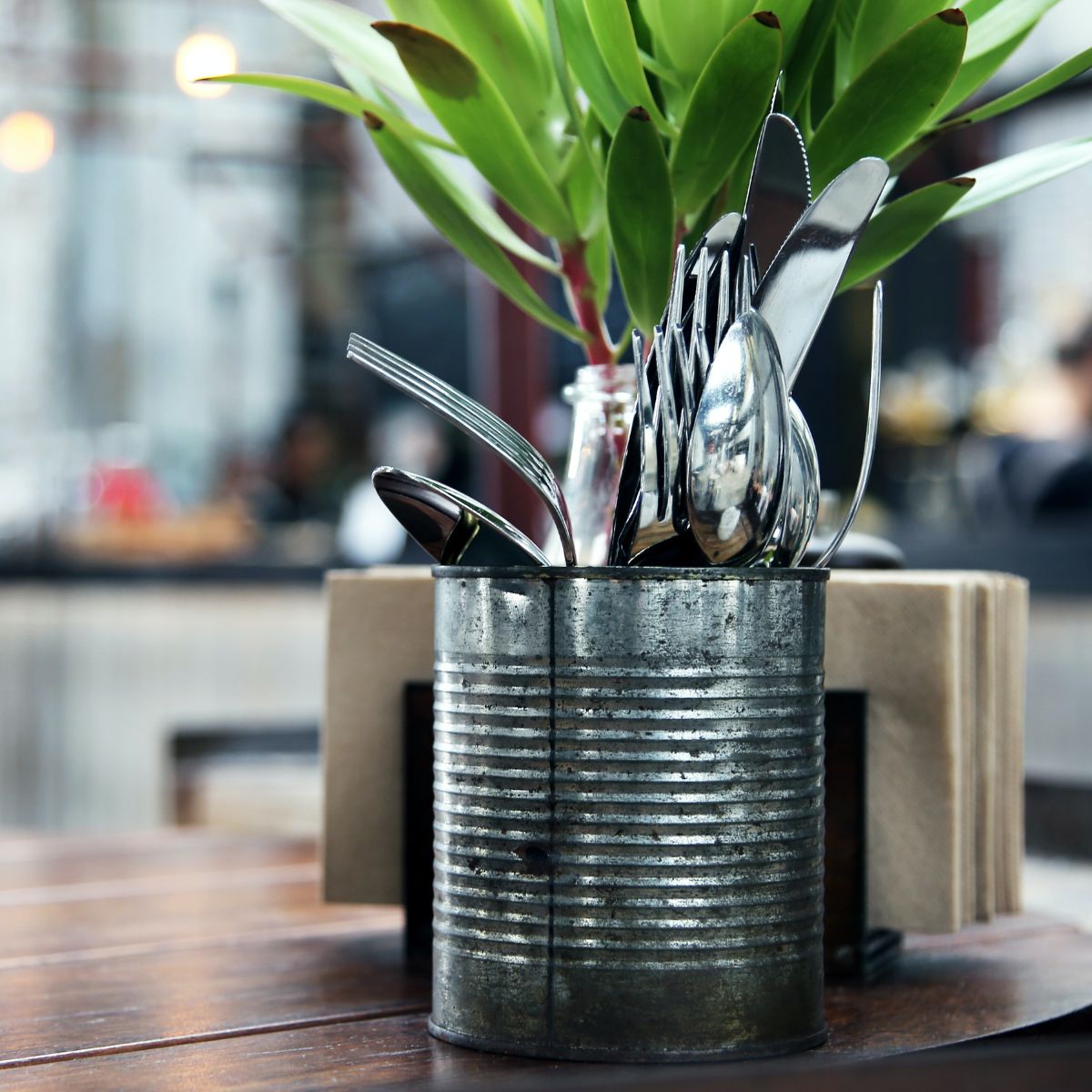 A metal can holds eating untensils on a table next to a plant.