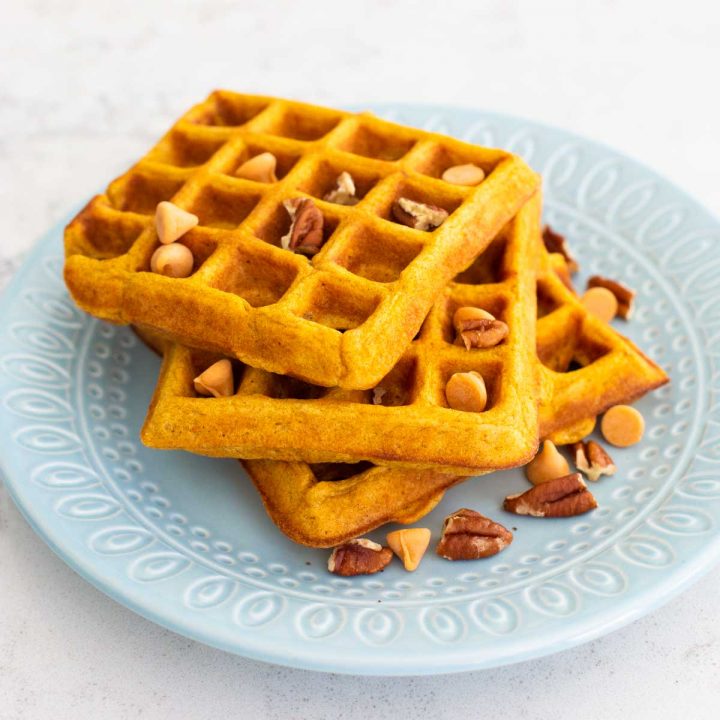 A blue plate has 3 pumpkin waffles sprinkled with butterscotch chips and pecans.