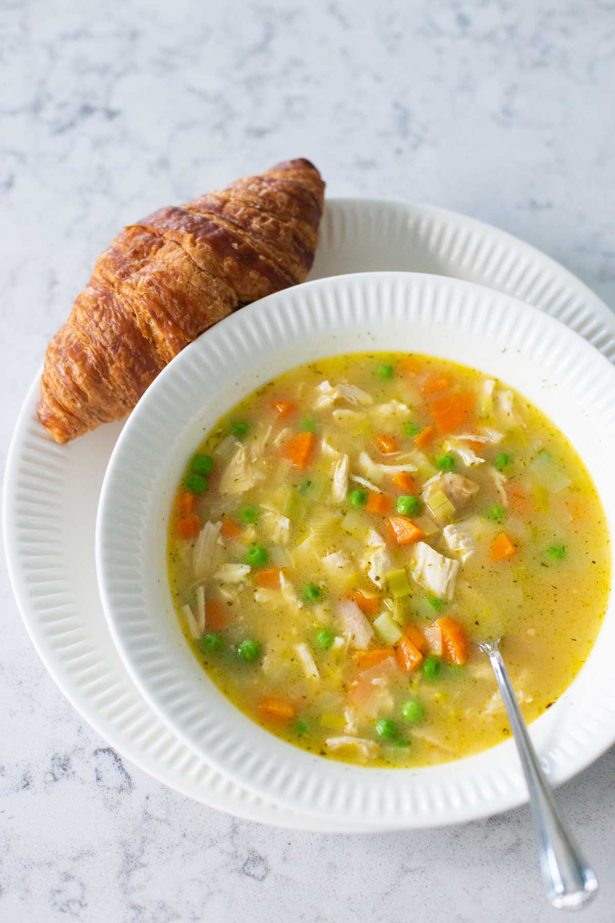 A bowl of chicken pot pie soup has a croissant on the plate next to it.