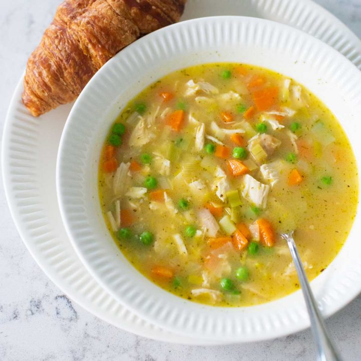 A bowl of chicken pot pie soup has a croissant on the plate next to it.