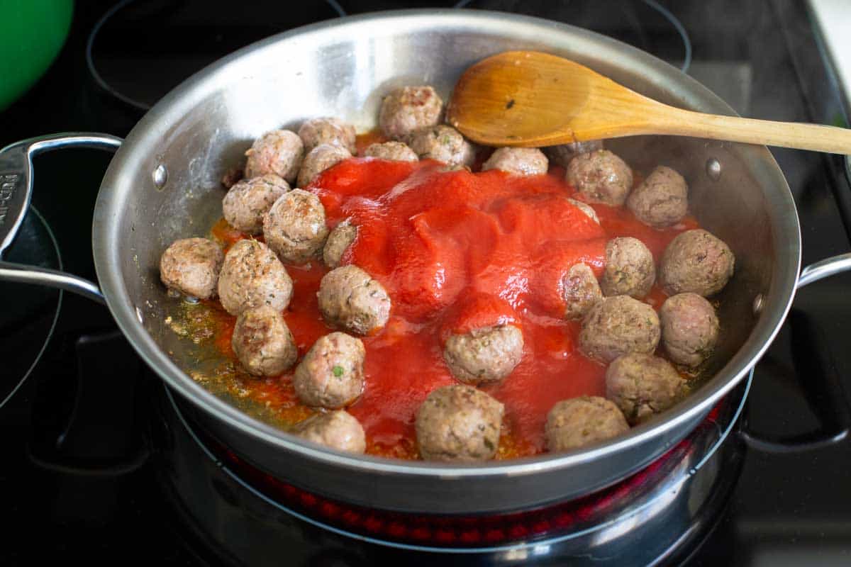 A wooden spoon is ready to scrape up the browned bits on the bottom of the pan using the tomato sauce to help deglaze.