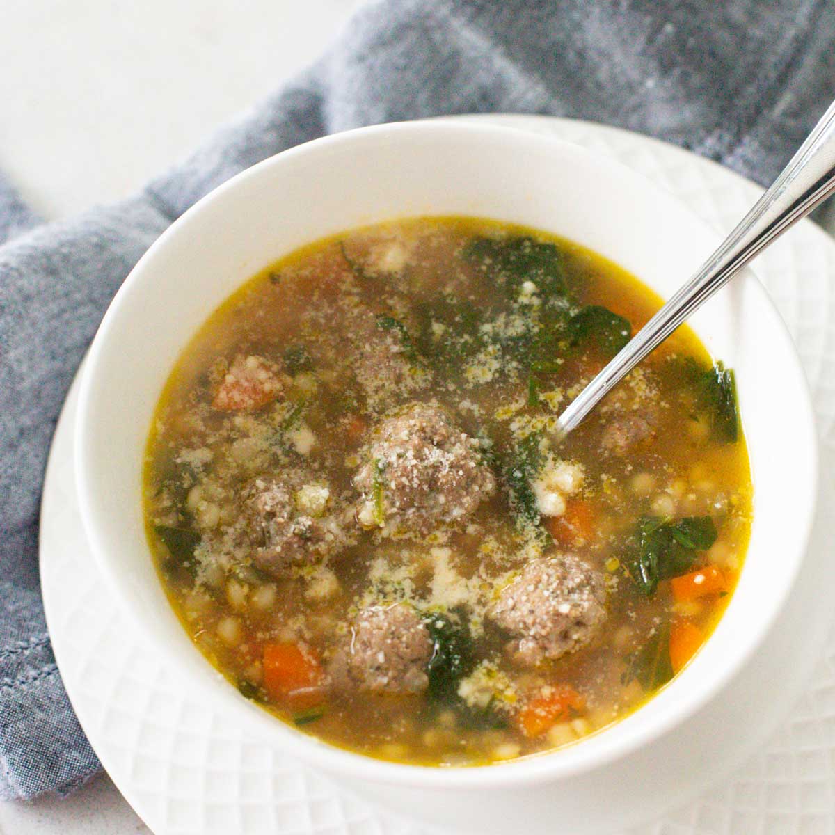 A bowl of soup has meatballs, couscous, and carrots with spinach in chicken broth.
