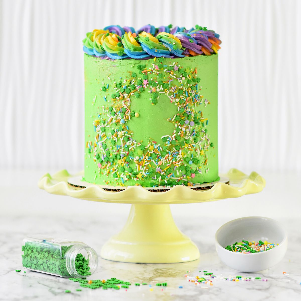 A green cake with rainbow frosting and green sprinkles on a cake stand.