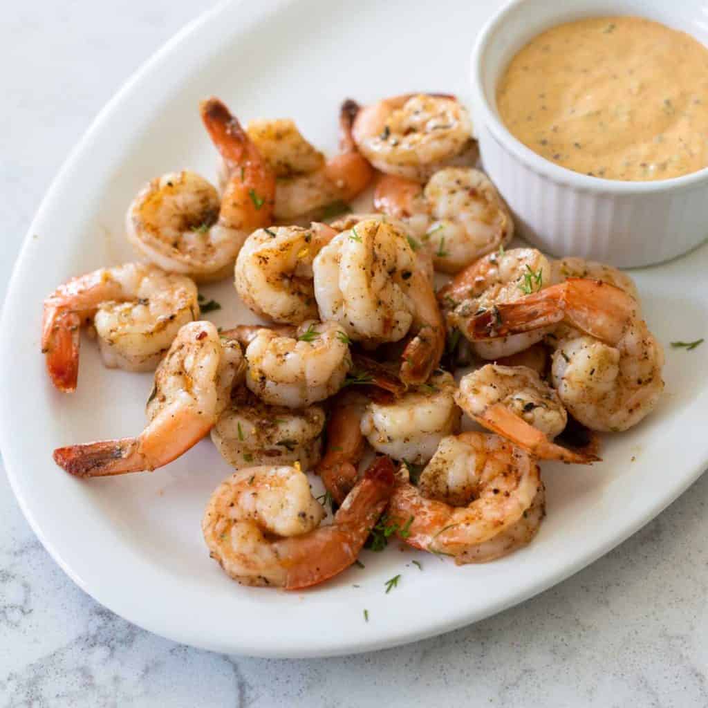 A platter of cooked shrimp next to a dipping cup of sauce for dunking.