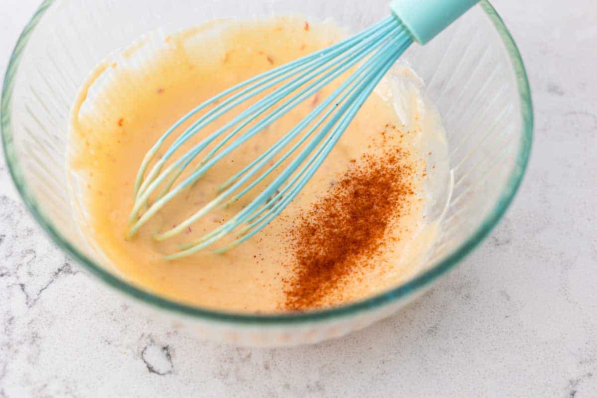 The bang bang sauce has been whisked together, cayenne powder is sprinkled on top. A blue whisk rests in the bowl.