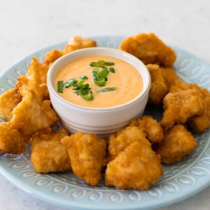 The bang bang sauce is in a dipping cup on a plate, surrounded by chicken nuggets.
