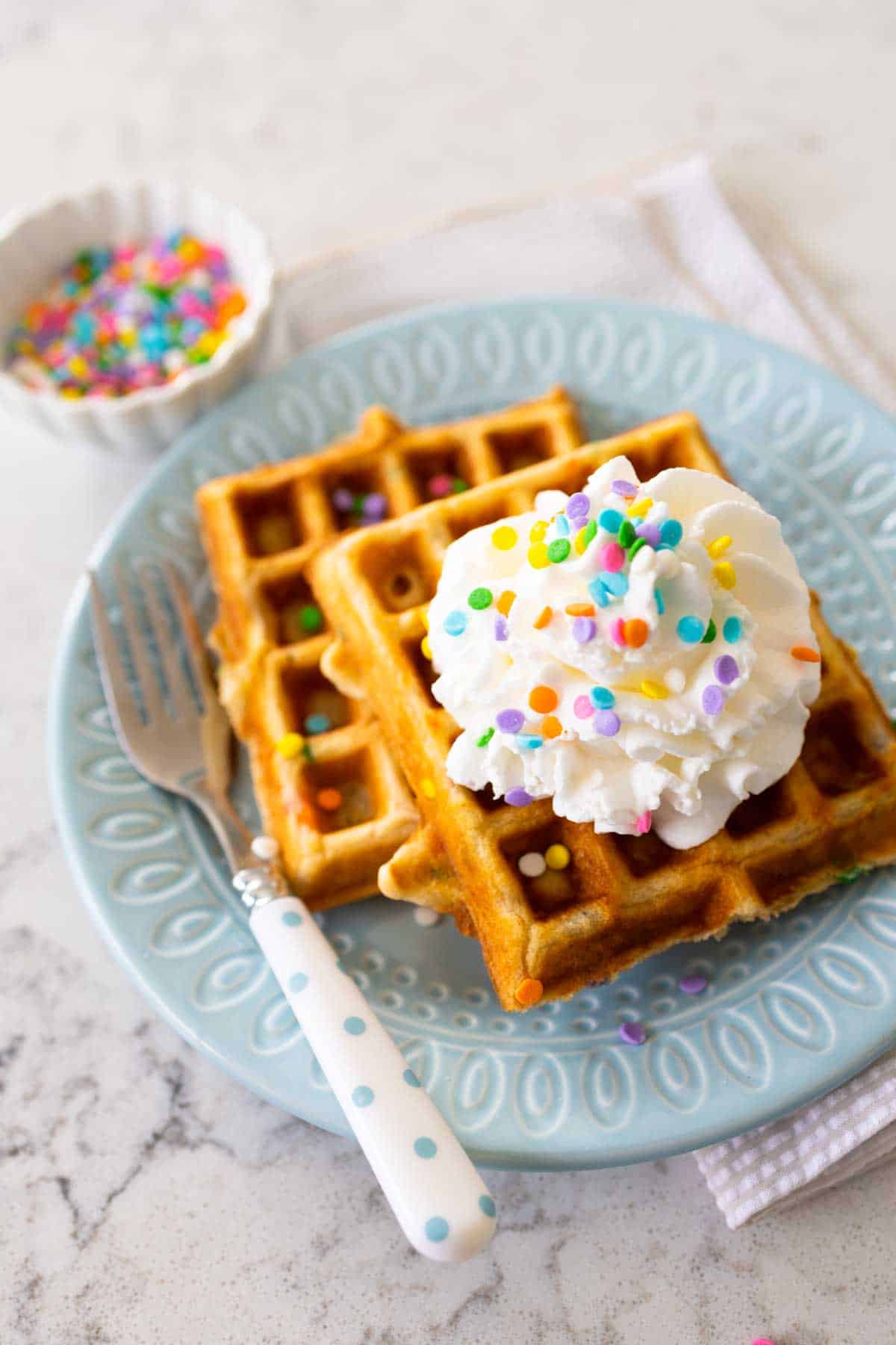 Two waffles on a blue plate have a dollop of whipped cream and pastel sprinkles.