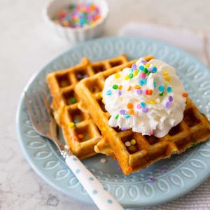 Two waffles on a blue plate have a dollop of whipped cream and pastel sprinkles.