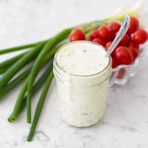 A mason jar filled with homemade buttermilk dressing sits next to a bunch of green onions and a bowl of cherry tomatoes.