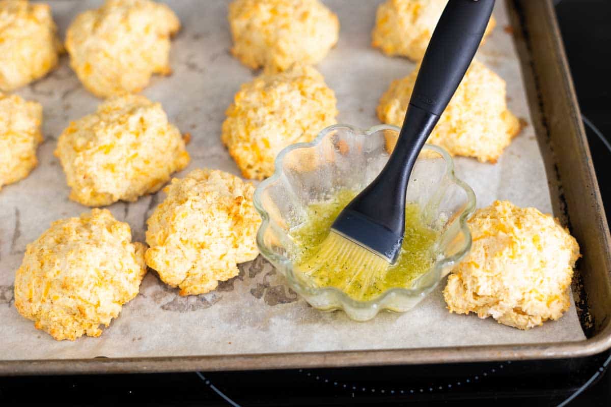 A pastry brush in a cup of melted butter is painting the baked biscuits.