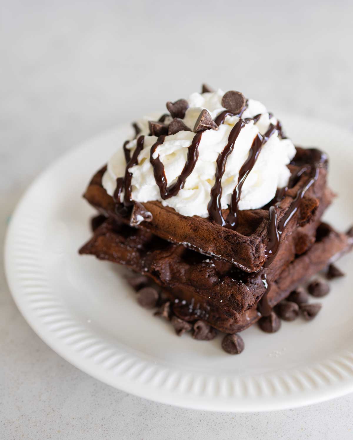 A stack of chocolate waffles with a large dollop of whipped cream, drizzle of chocolate sauce, and sprinkles of chocolate chips on a white plate.