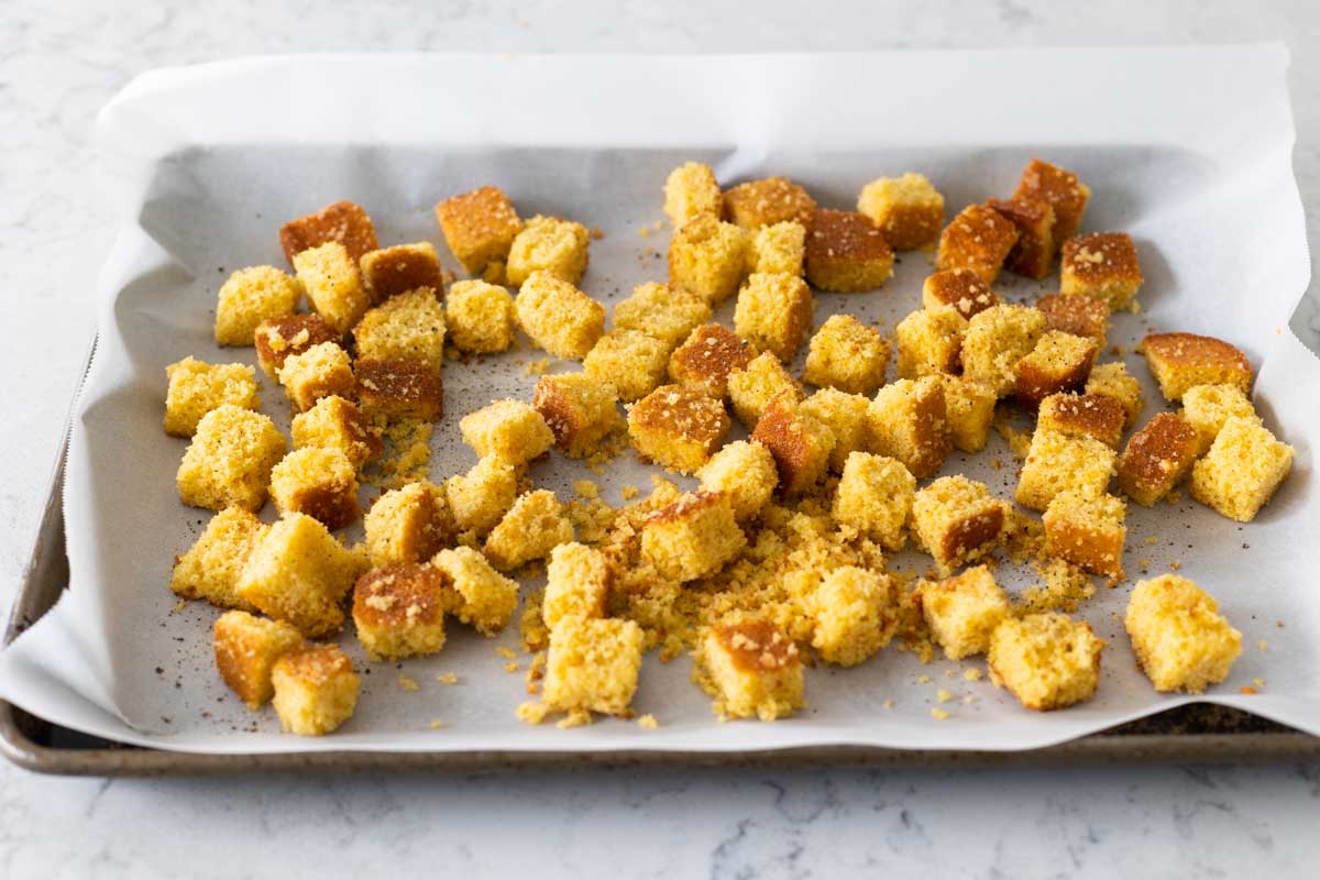 The cubes of cornbread have been put on parchment paper on a baking sheet for baking.