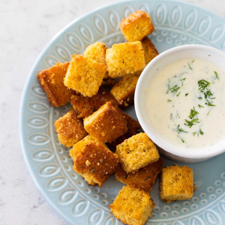 Homemade cornbread croutons on a blue plate next to a bowl of buttermilk dressing.