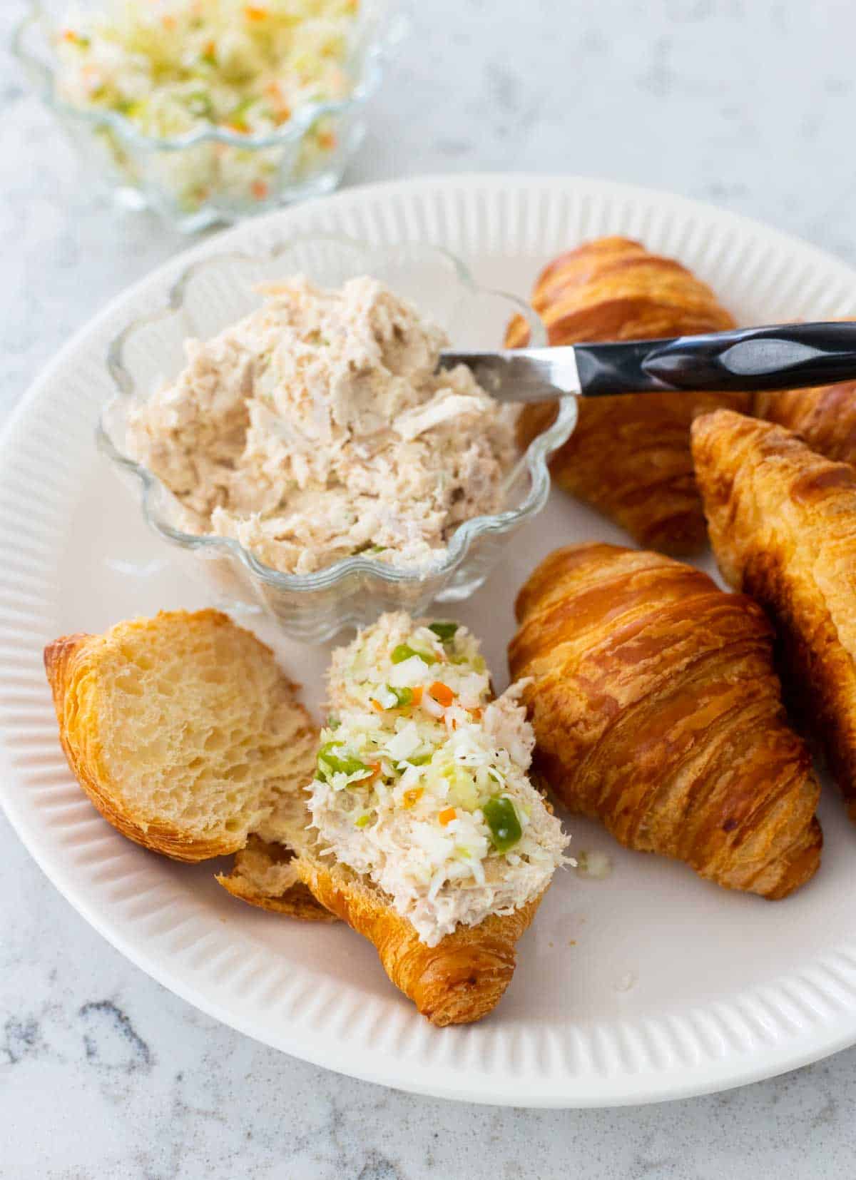 A plate filled with mini croissants, a bowl of chicken salad, and a container of slaw. One sandwich is being prepped.