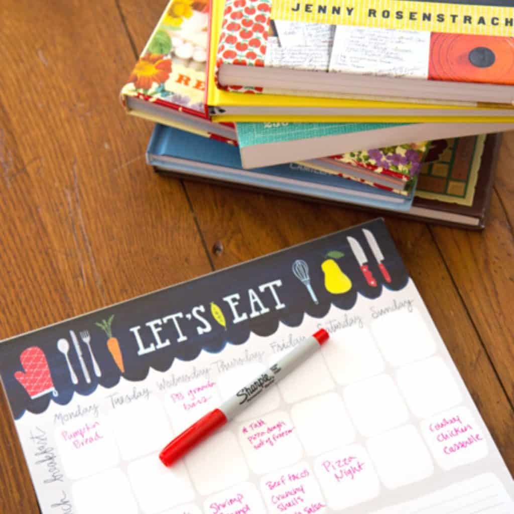 A meal planning pad with pen sits next to a stack of cookbooks.