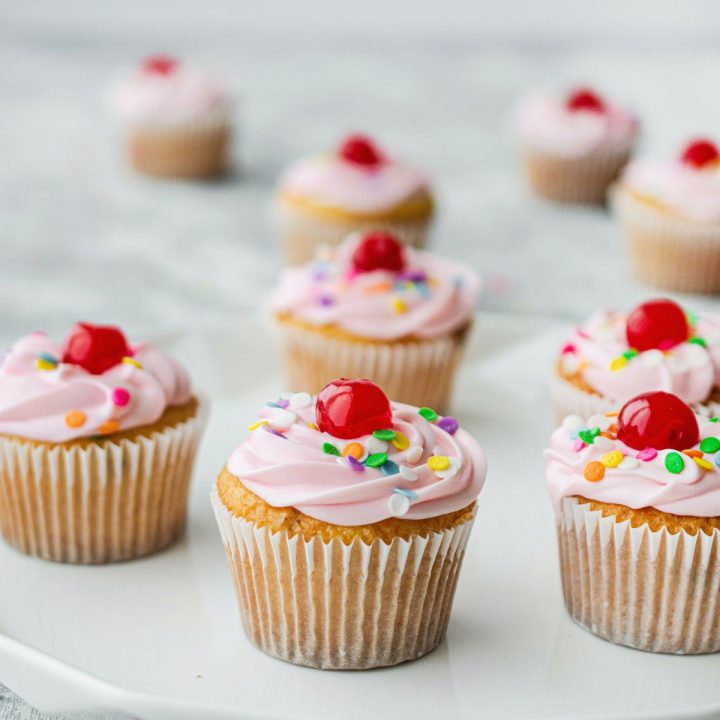 Pink frosted cupcakes with a red cherry on top and sprinkles.