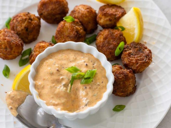 The remoulade sauce is on a plate with mini crab cakes and a spoon. Lemon wedges are scattered around and green onions sprinkled on top.
