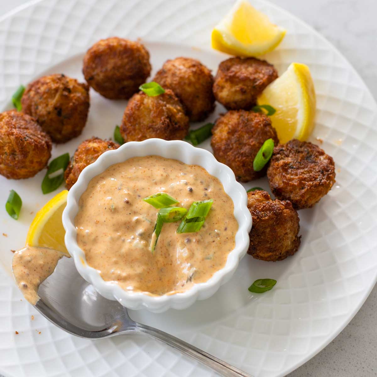 The remoulade sauce is on a plate with mini crab cakes and a spoon. Lemon wedges are scattered around and green onions sprinkled on top.