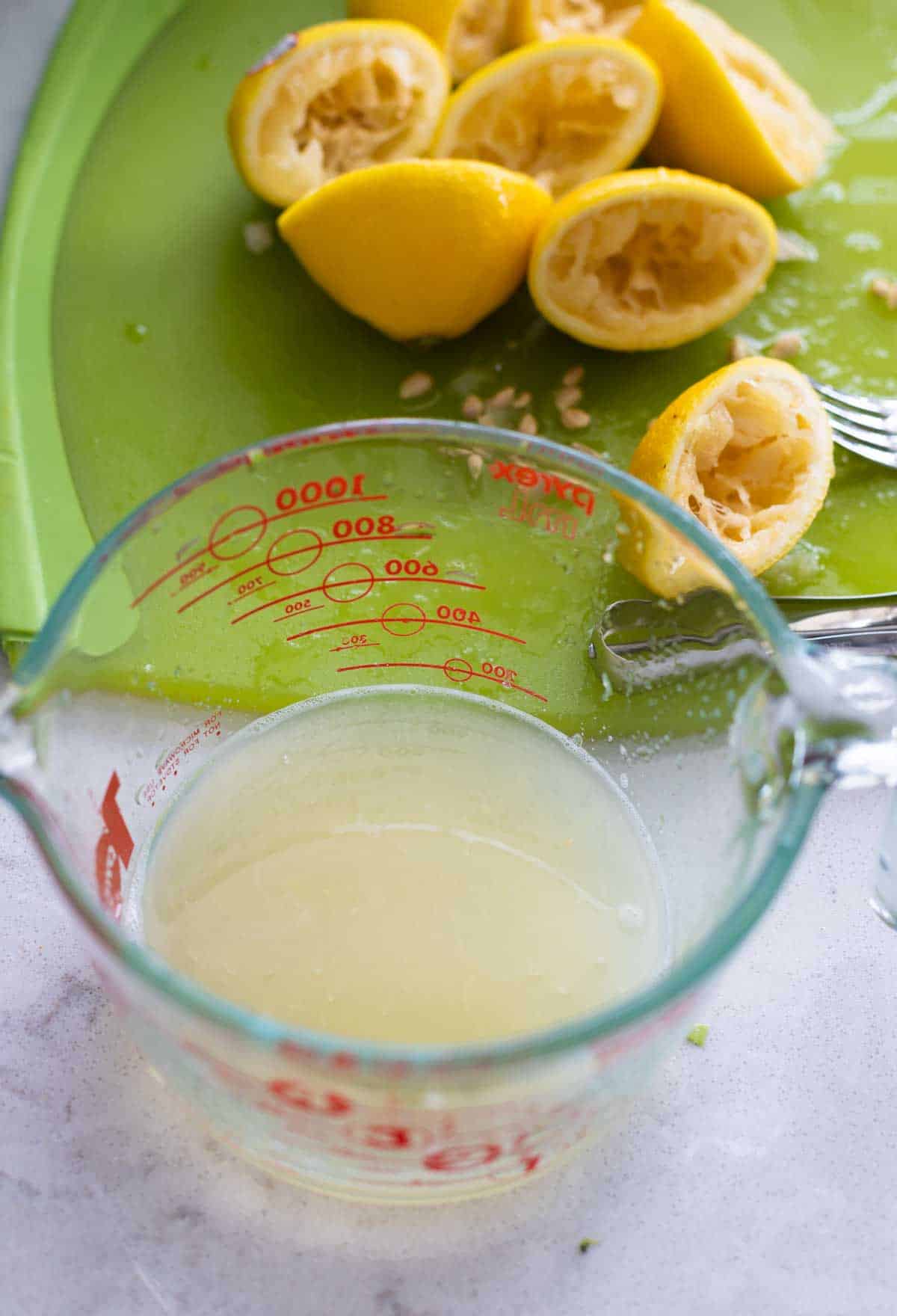 A measuring cup of lemon juice next to a pile of squeezed lemons.