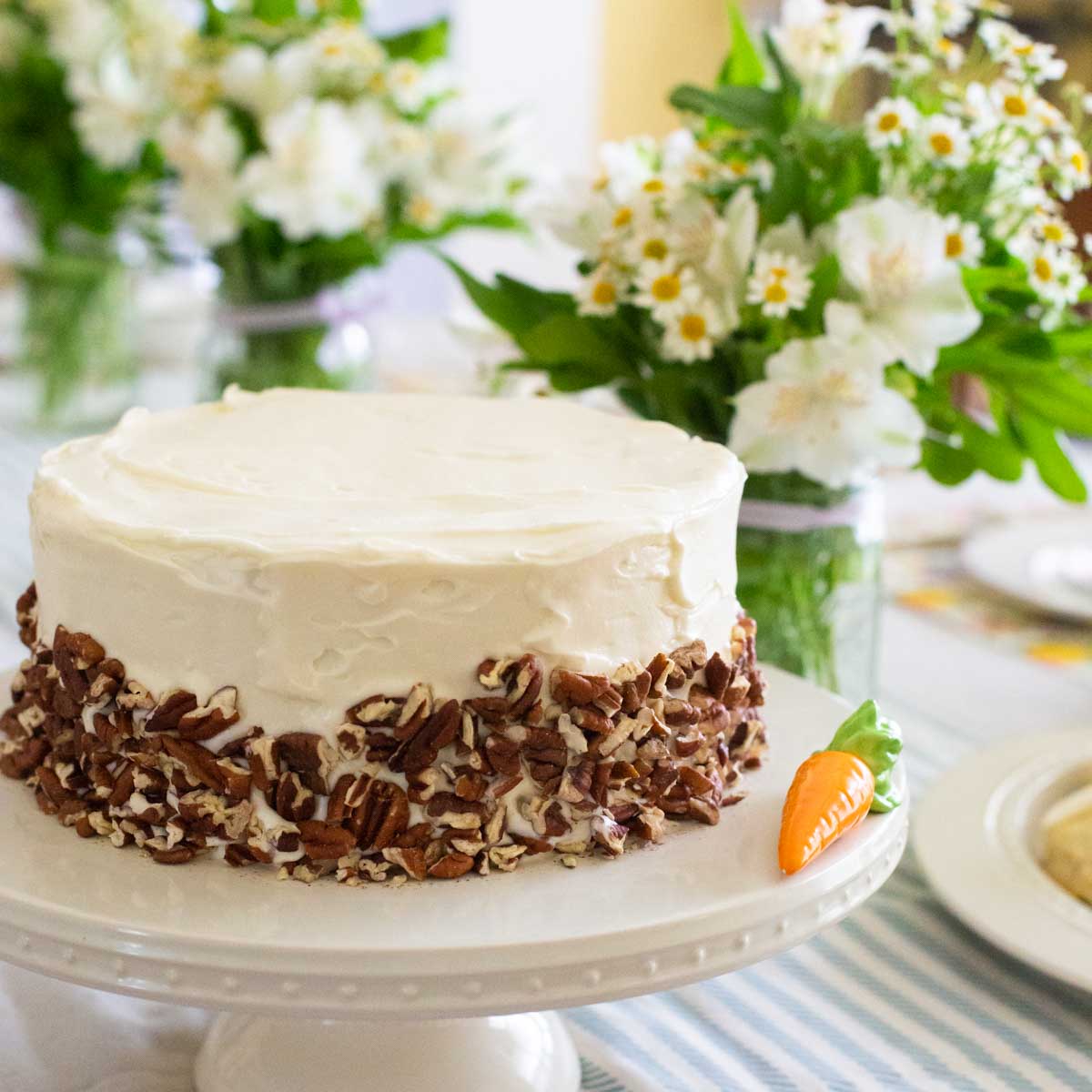 A gorgeous carrot cake with pecans on a cake platter next to bouquets of fresh flowers.
