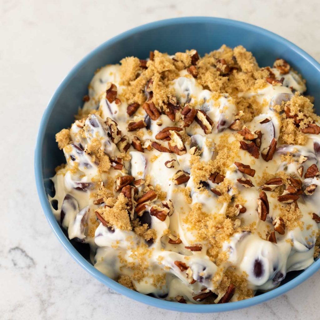 A big blue bowl is filled with a creamy grape salad with cream cheese, covered in brown sugar and pecans.