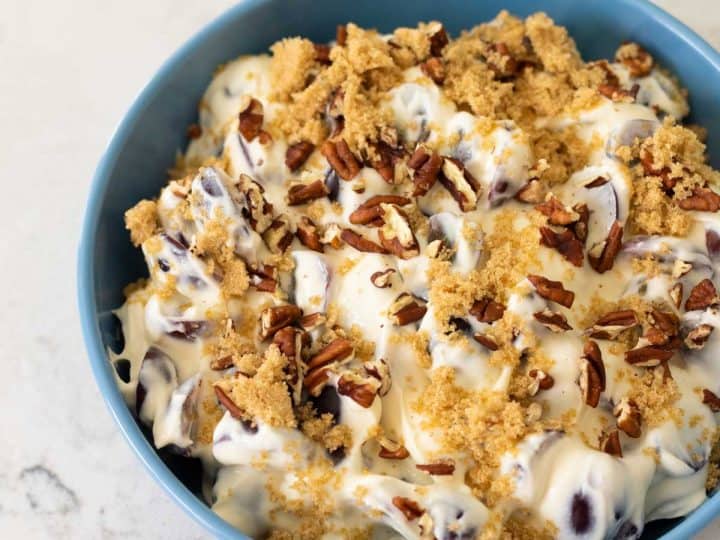 A big blue bowl is filled with a creamy grape salad with cream cheese, covered in brown sugar and pecans.