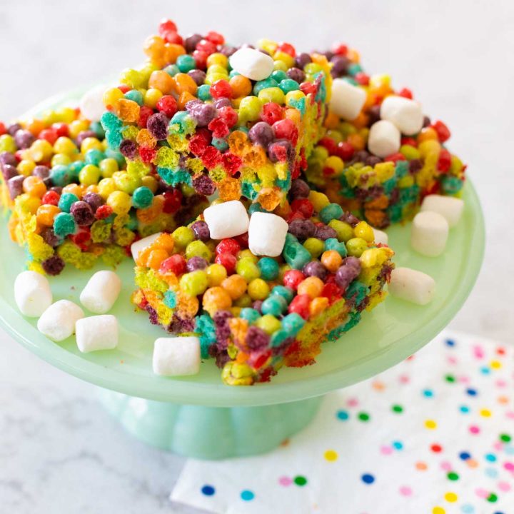 A cake plate has several squares of cereal bars in rainbow colors with mini marshmallows scattered about.
