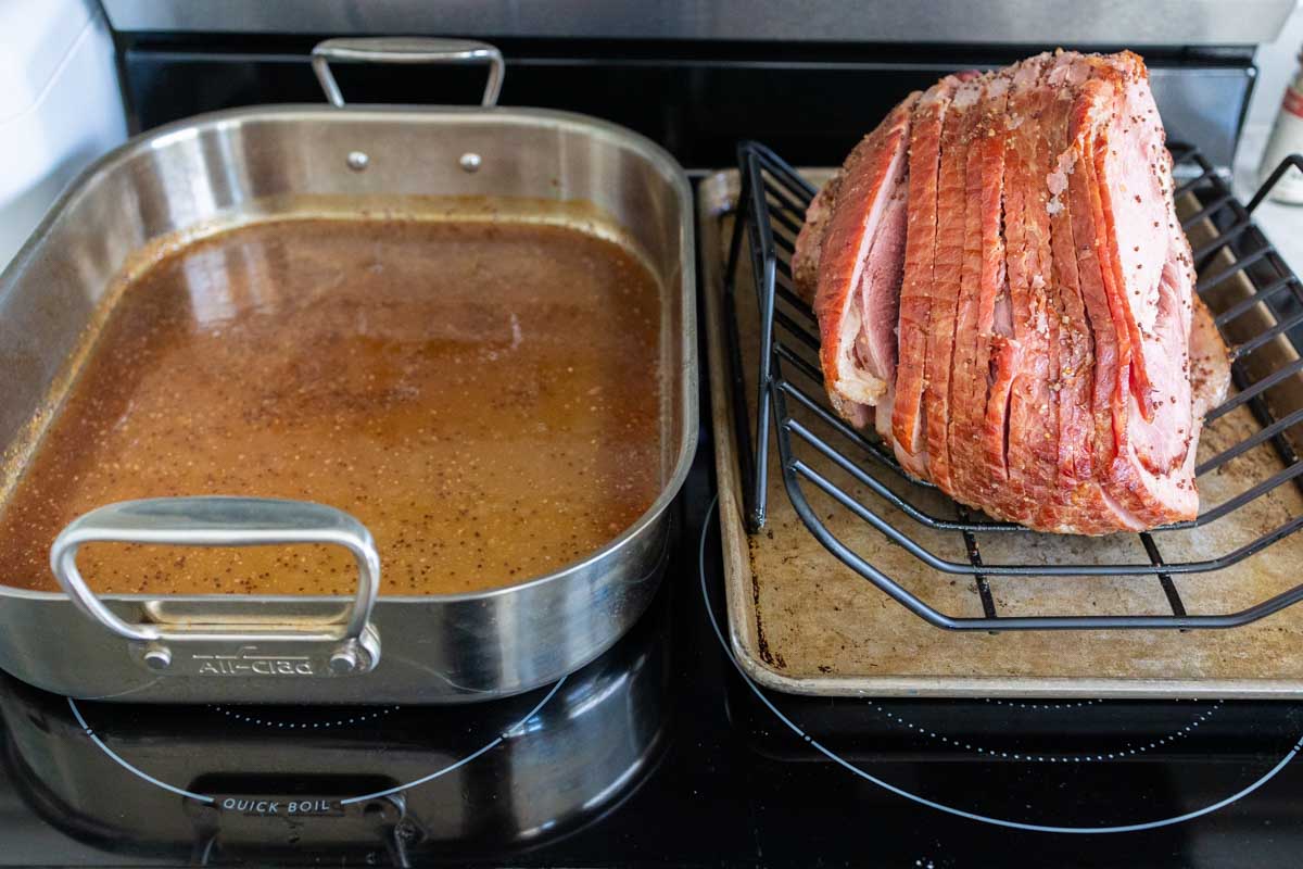 The ham is resting on a rack next to the roasting pan which is filled with glaze.