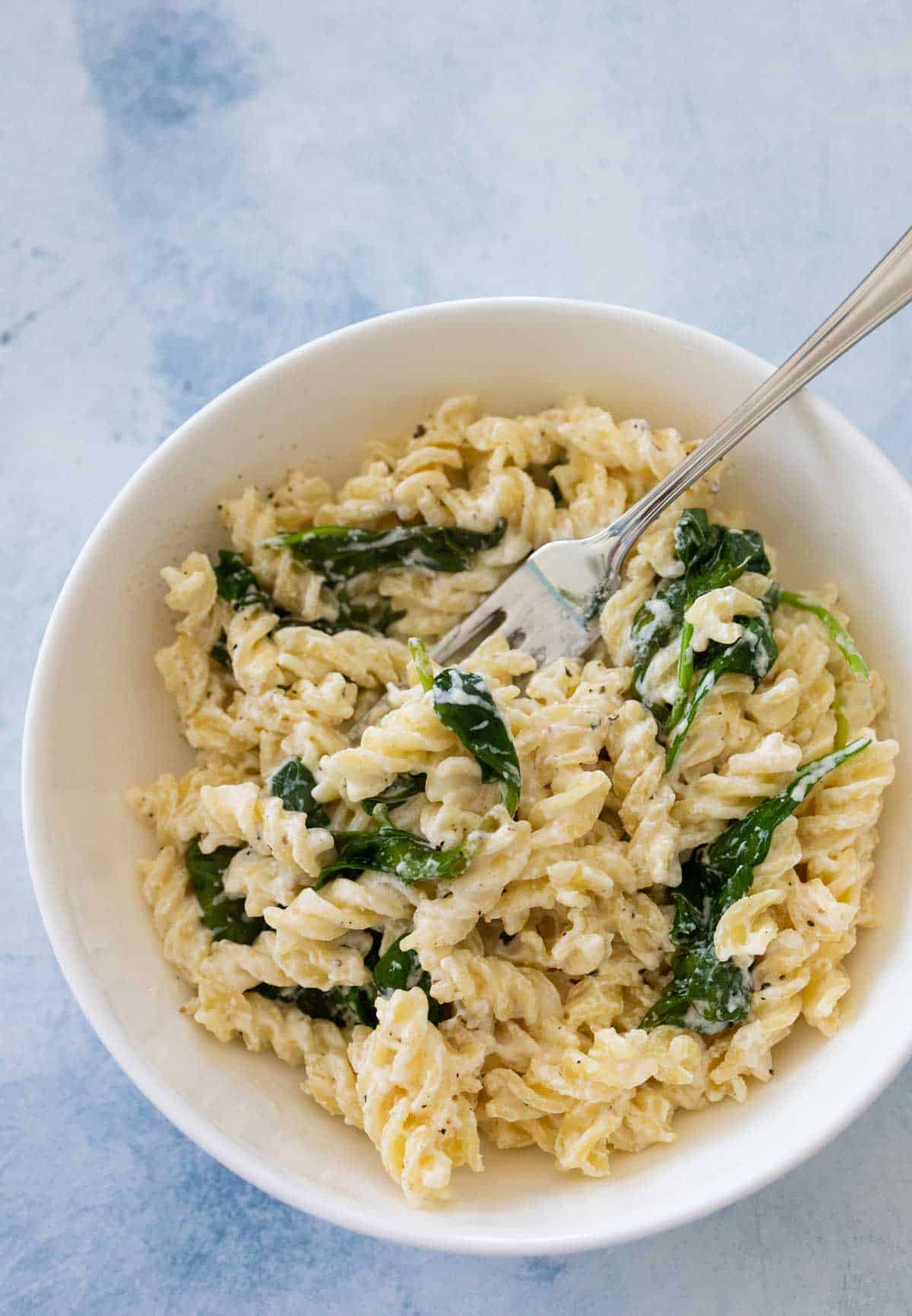 A white bowl with a fork has a serving of goat cheese pasta.