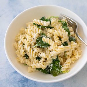 A white bowl filled with creamy spiral noodles with baby spinach leaves mixed in.