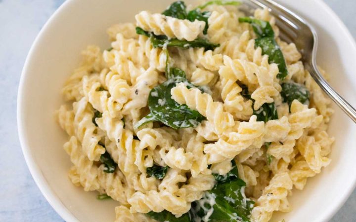 A white bowl filled with creamy spiral noodles with baby spinach leaves mixed in.