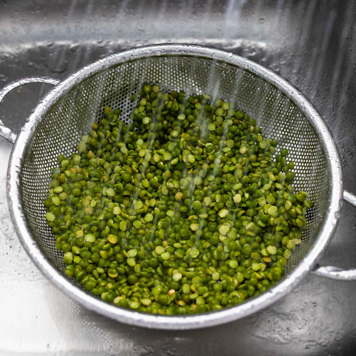 The peas are in a strainer in the sink. Water is rinsing them off.