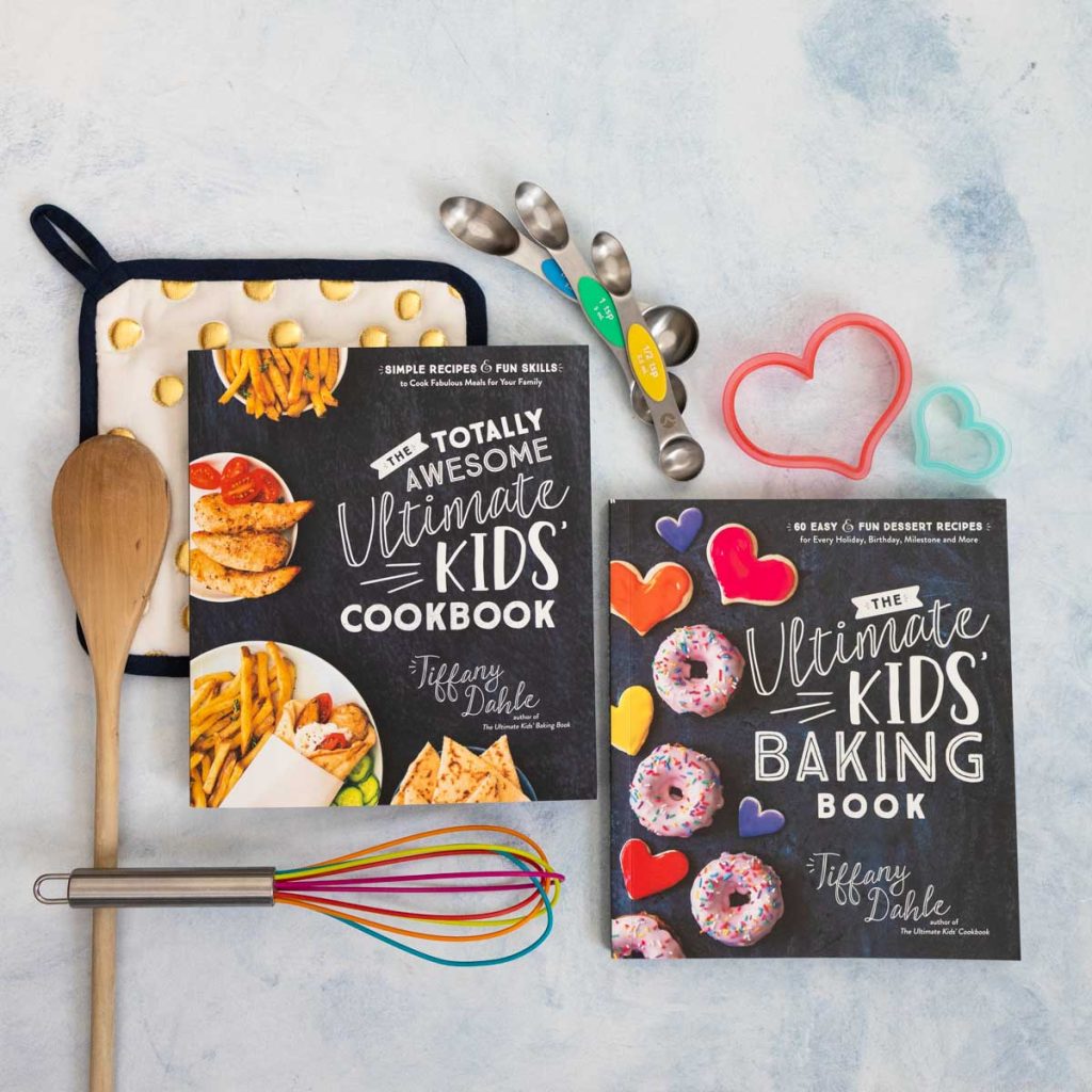 Copies of The Totally Awesome Ultimate Kids' Cookbook and The Ultimate Kids' Baking Book by Tiffany Dahle sit next to a wooden spoon, a rainbow whisk, a pot holder, measuring spoons, and heart-shaped cookie cutters.