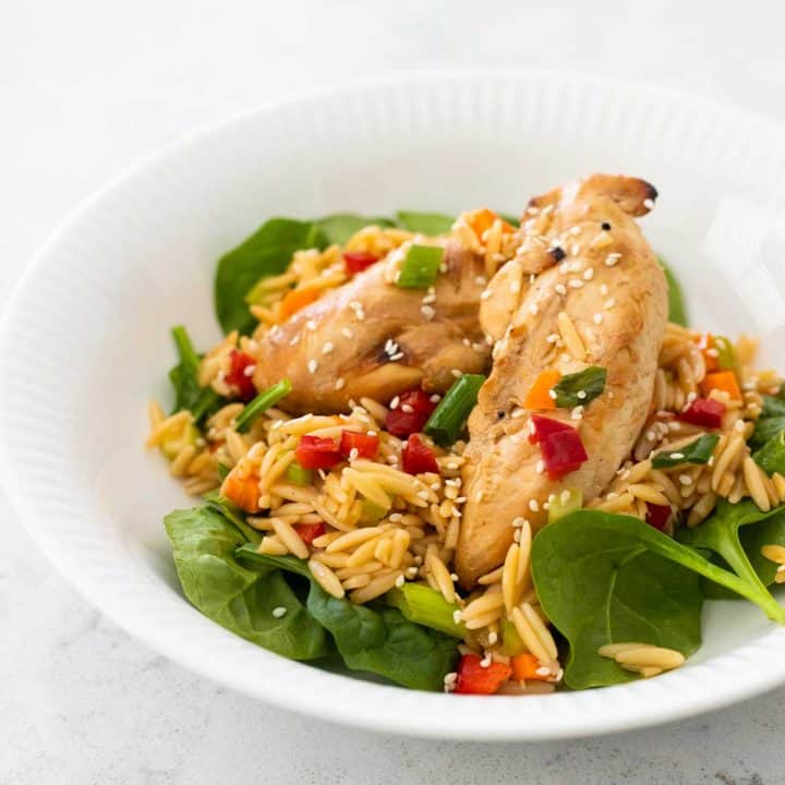 The finished salad is in a white bowl with 2 chicken tenderloins on a bed of pasta salad with baby spinach.