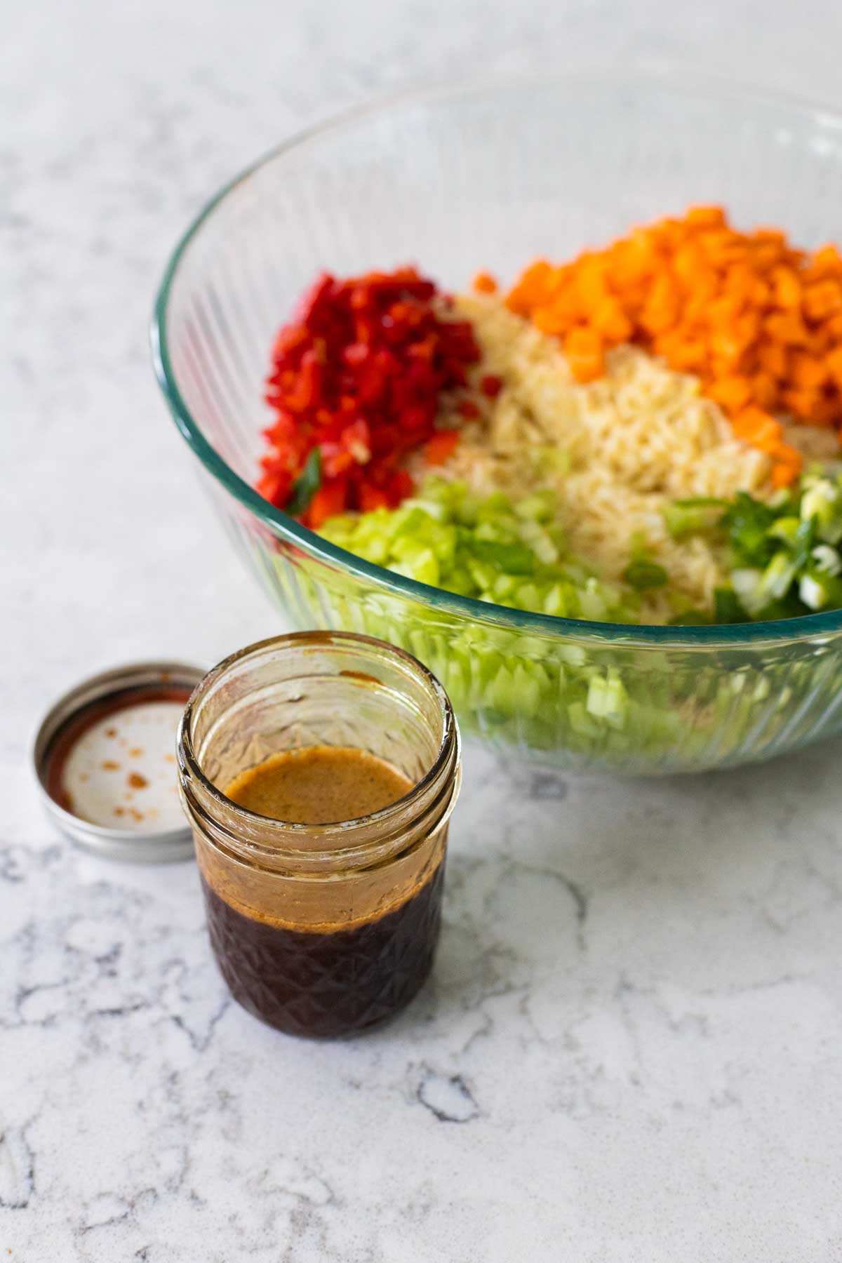 The mason jar of Asian marinade sits next to a mixing bowl filled with pasta salad and raw veggies.