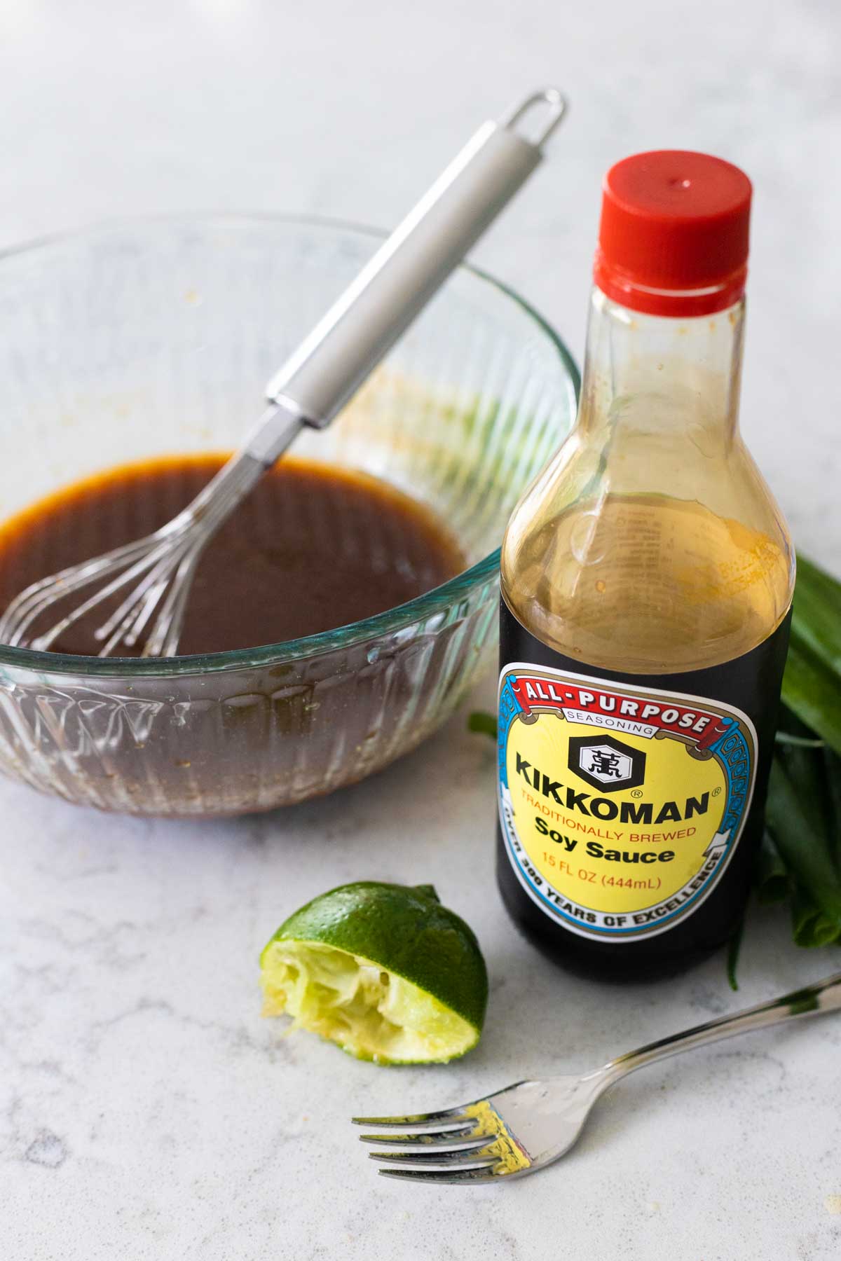 The mixing bowl has a whisk stirring the marinade. A bottle of soy sauce and half of a fresh lime with a fork sit next to the bowl.
