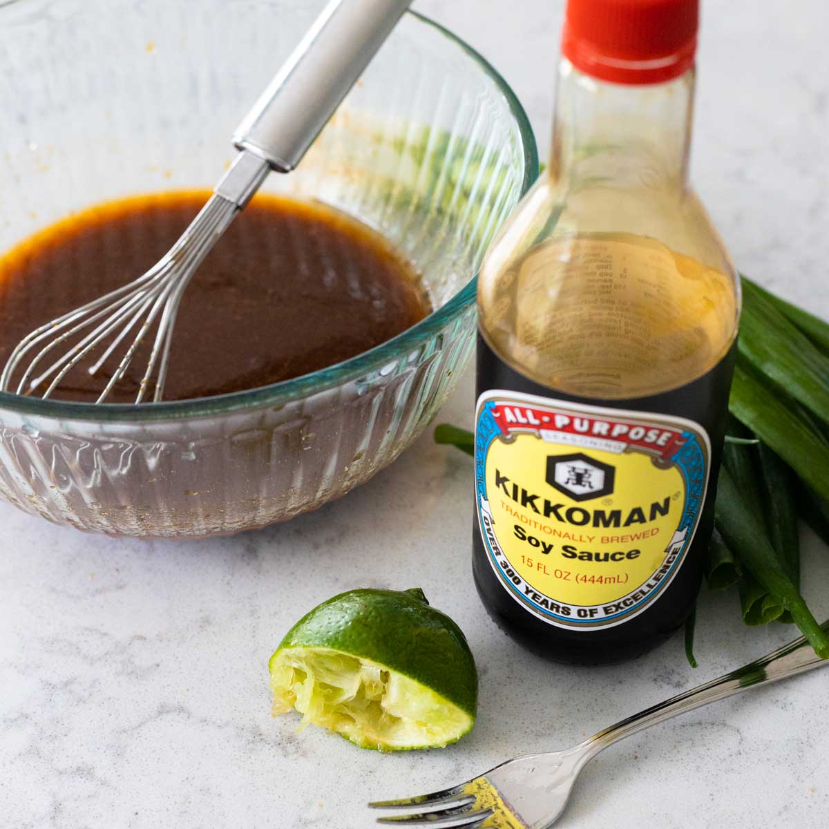 A mixing bowl with a whisk is mixing together the soy sauce, lime juice, and other ingredients for the marinade. A bottle of Soy sauce and a squeezed lemon are in front.